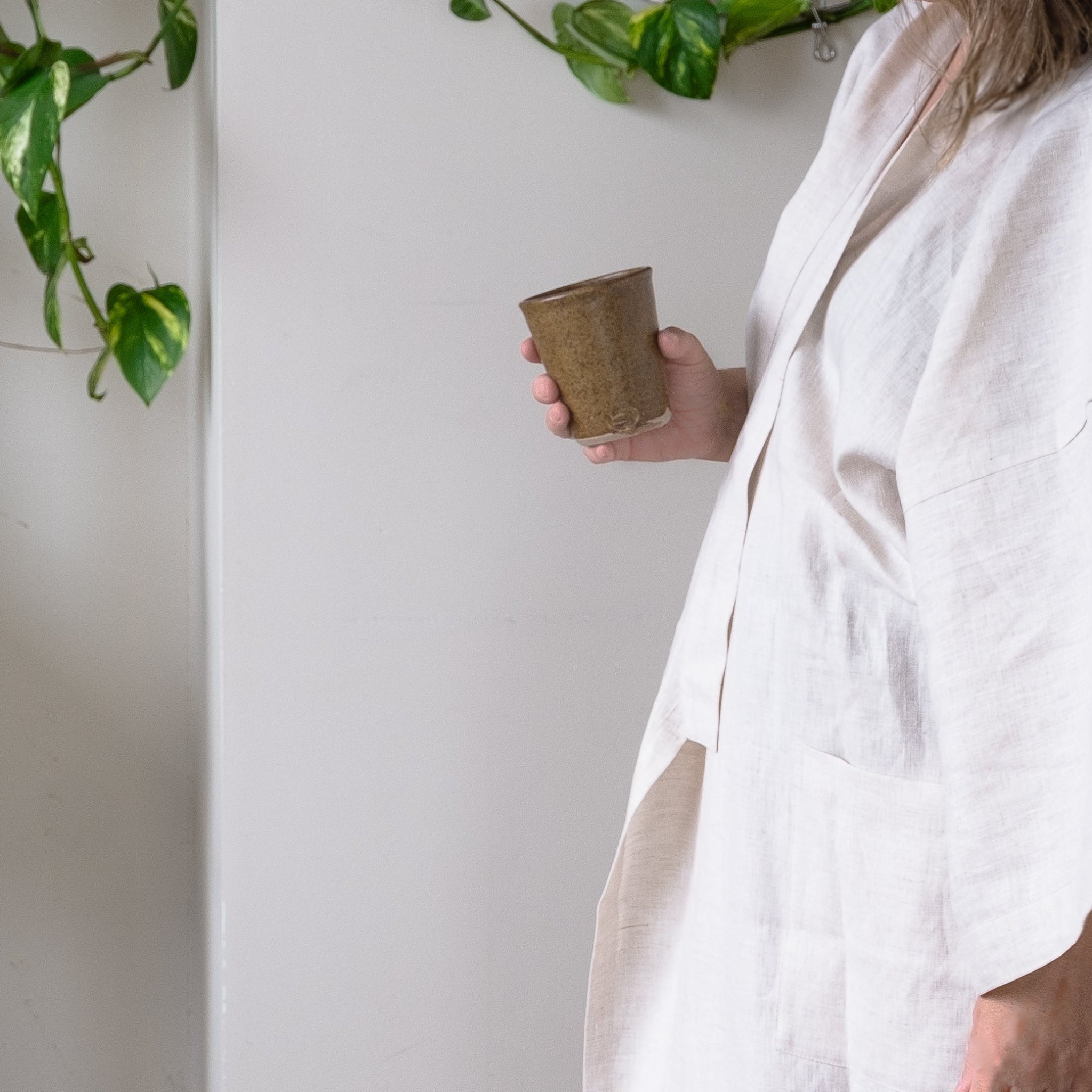 Person wearing an oat linen robe holding a small brown ceramic cup against a white wall with plants.