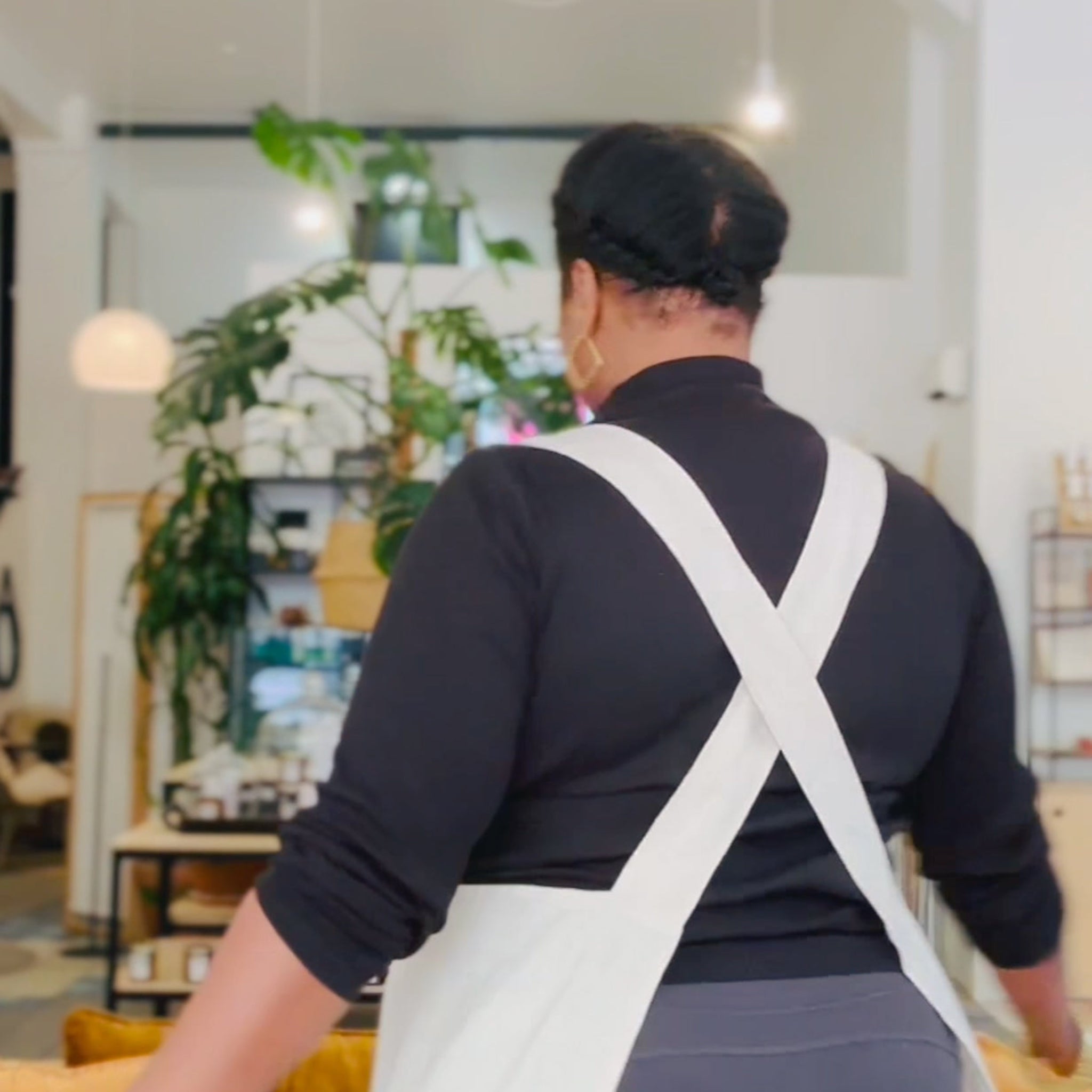 Person wearing a white apron in an indoor setting with plants and furniture.