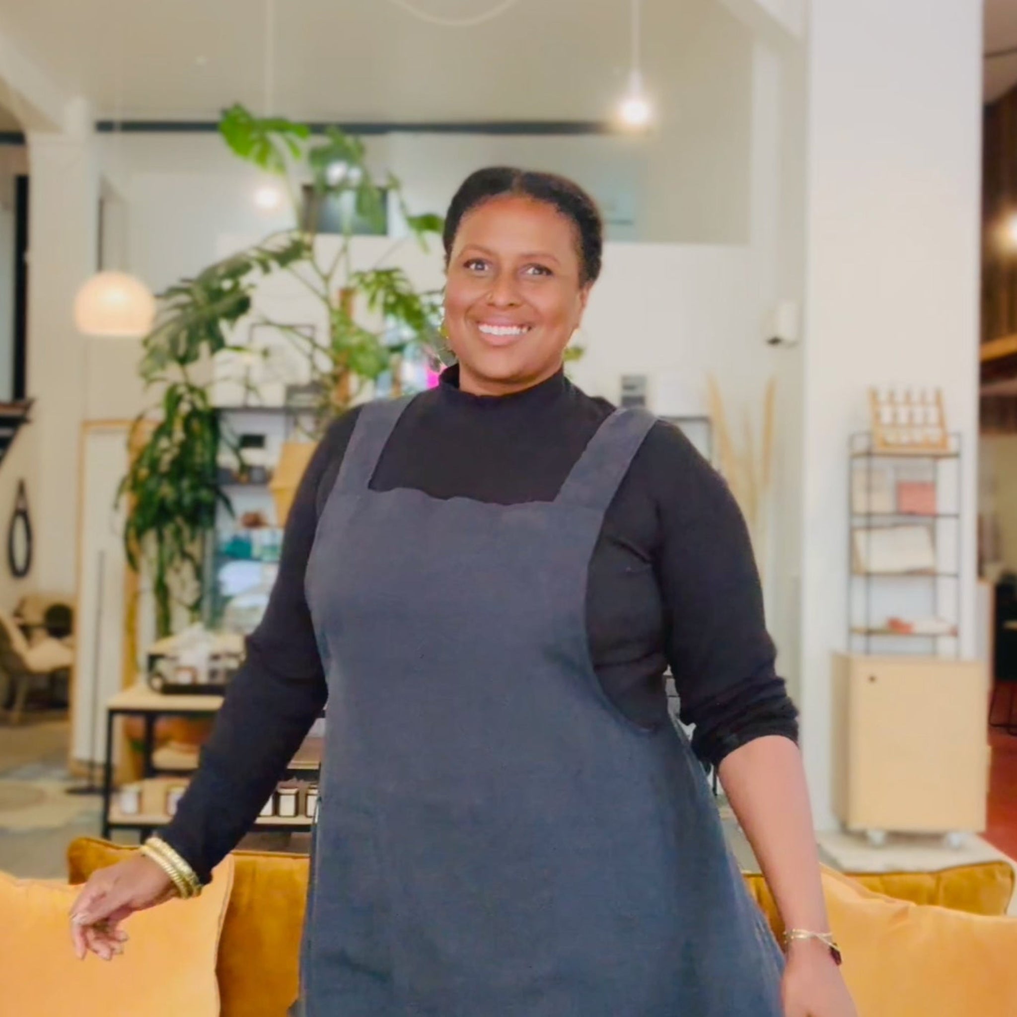 Woman wearing a charcoal black apron standing in a modern indoor setting with plants and furniture.