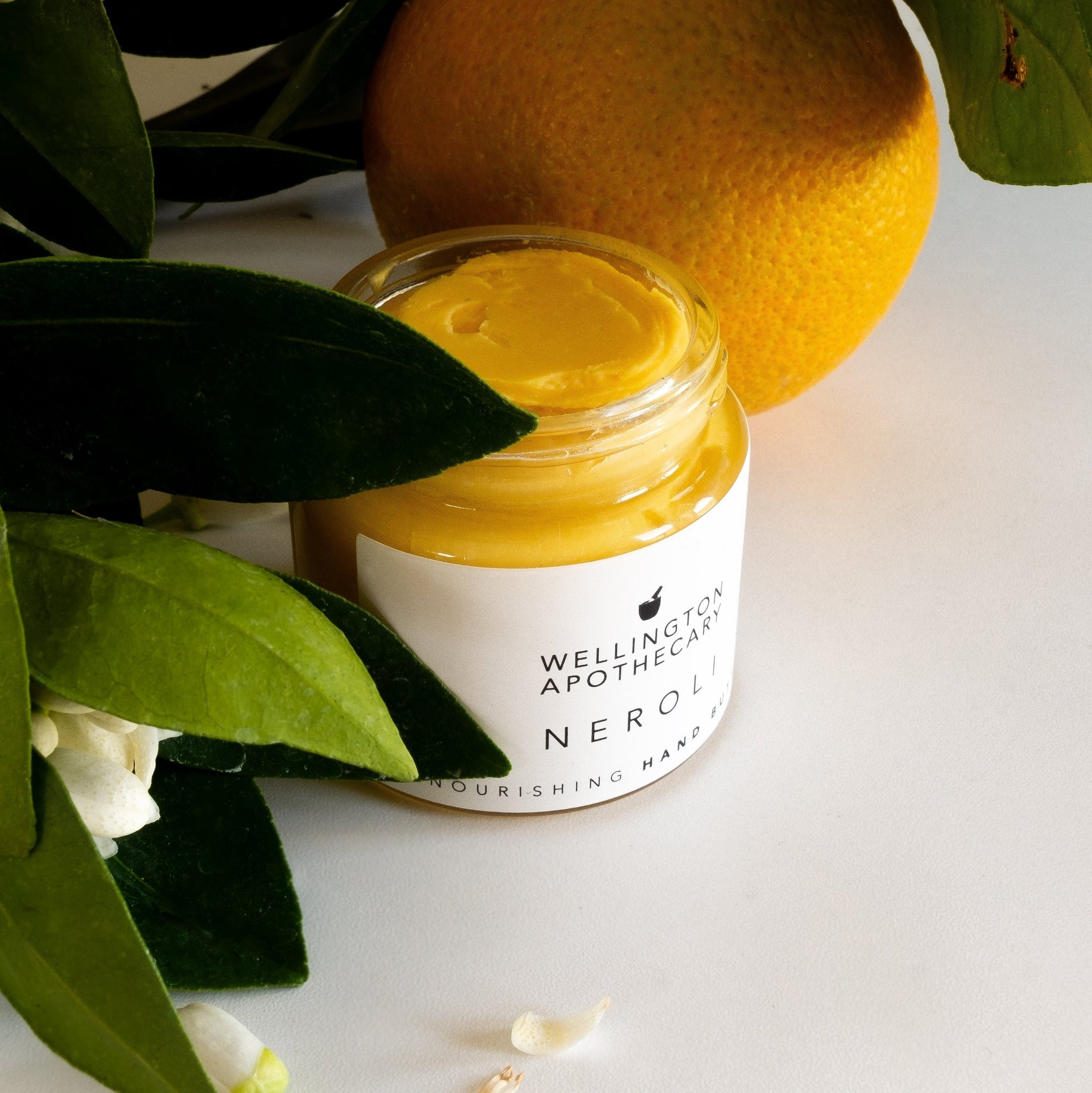 An open glass jar with mango yellow hand butter inside, surrounded by neroli leaves, flowers and fruit.