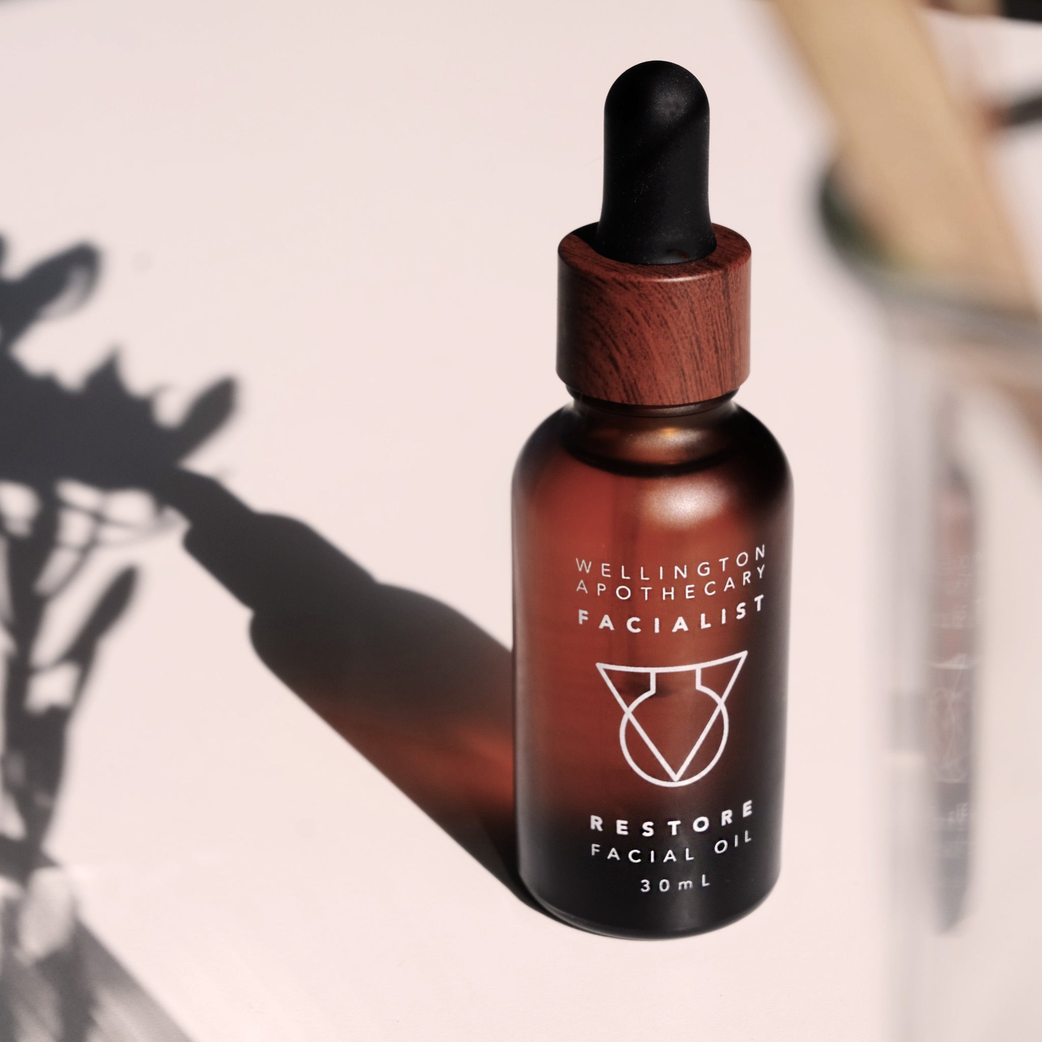 Amber glass bottle of restore facial oil on white bench with blurred beaker beside it and botanical shadows cast behind it.