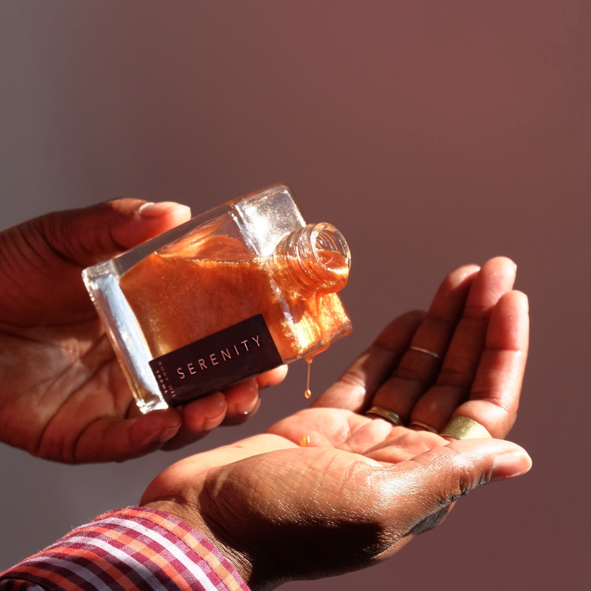 Hand pouring shimmering body oil from a bottle labeled &