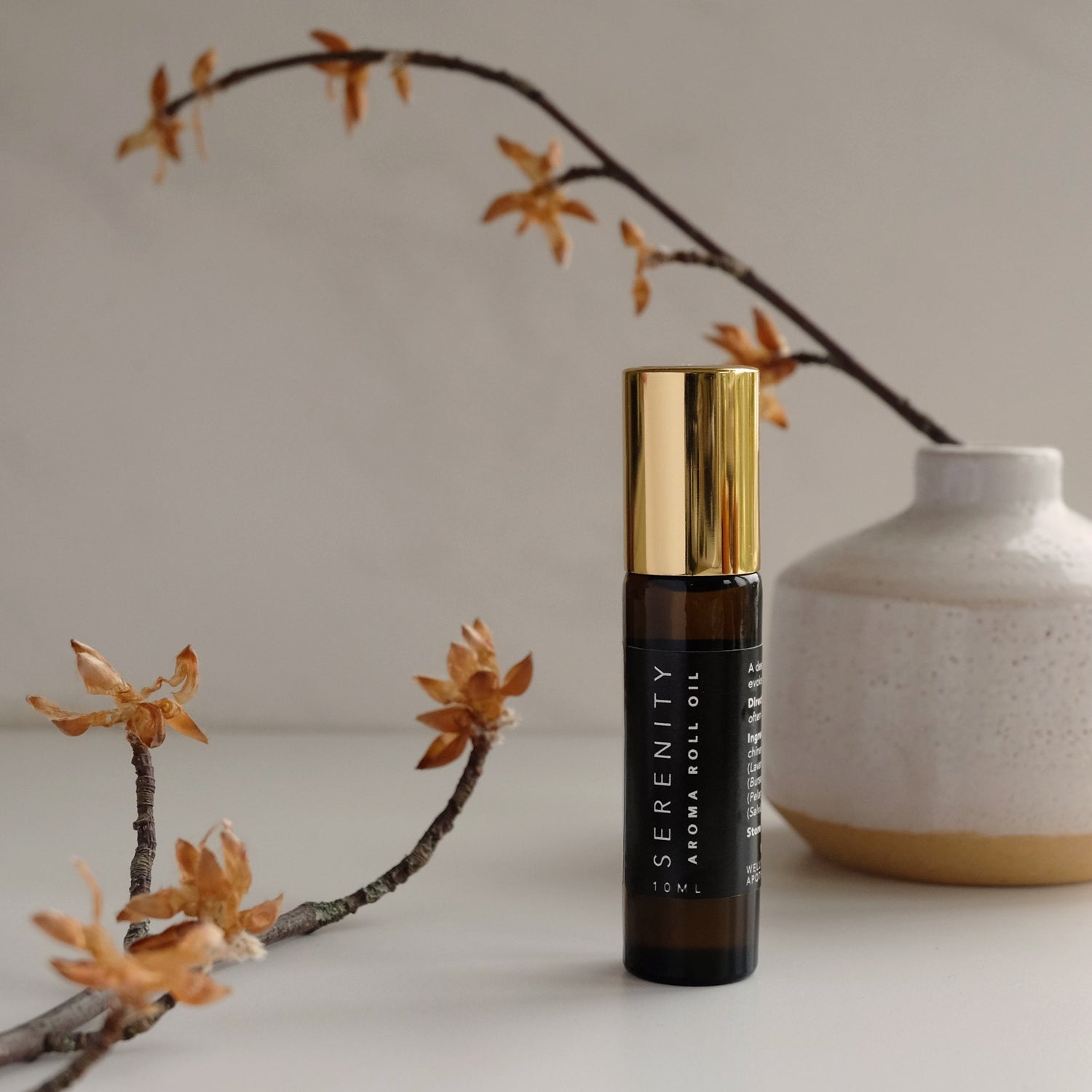 An amber glass vial of serenity perfume blend with gold lid, autumnal twigs and an earthy white ceramic vase surrounding.