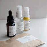 Four products on white bench including cleansing oil, toner spray, serum and clay mask in sachet.