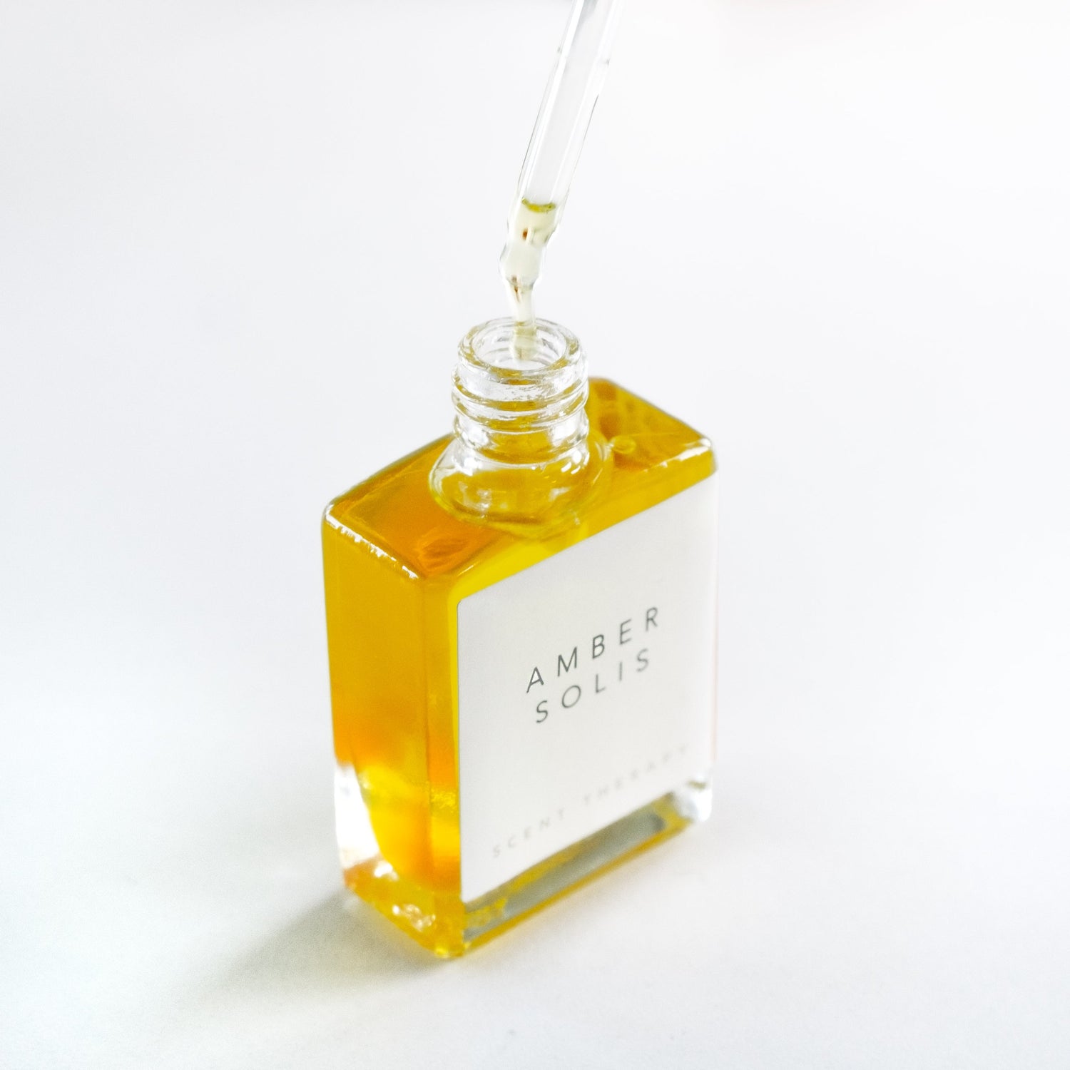 A square glass bottle of the Amber Solis oil with the dropper top removed and the golden oil dripping from it.