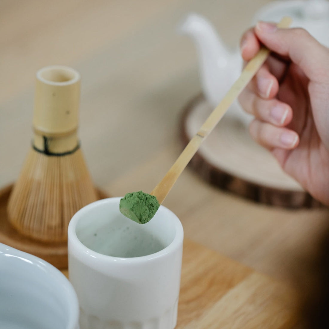 Bamboo match spoon with a scoop of bright green matcha powder from white ceramic vessel and other matcha tea accessories surrounding.