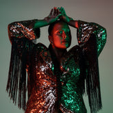 Portrait of Deva in dark light wearing a shimmering pink and green jacket with long black tassels.