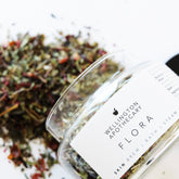 A glass apothecary jar tipped over spilling out the dried herbal tea blend of Flora Skin Brew.