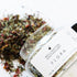 A glass apothecary jar tipped over spilling out the dried herbal tea blend of Flora Skin Brew.
