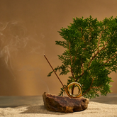 Hinoki incense burner in brass holder a top a rock and sand with conifer branch behind.