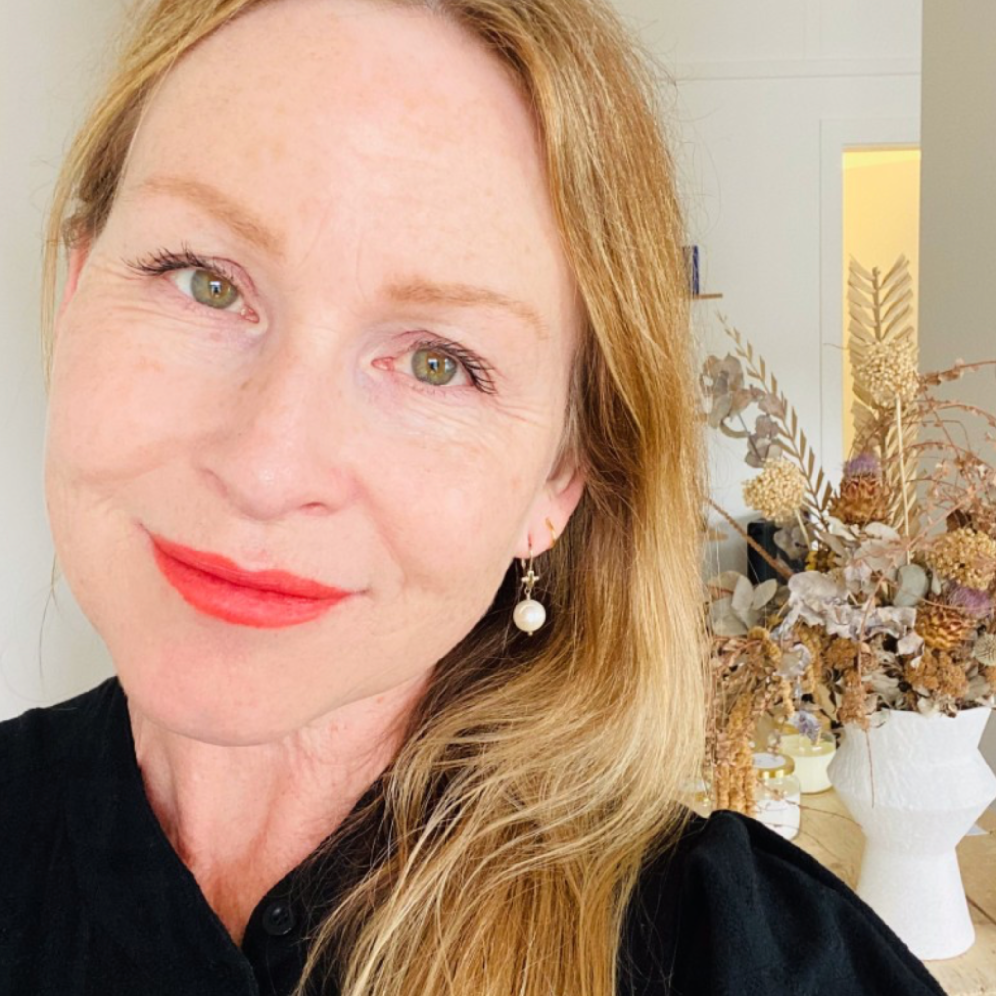 Portrait of Mel wearing red lipstick and pearl earrings, dried flower bouquet in the background.