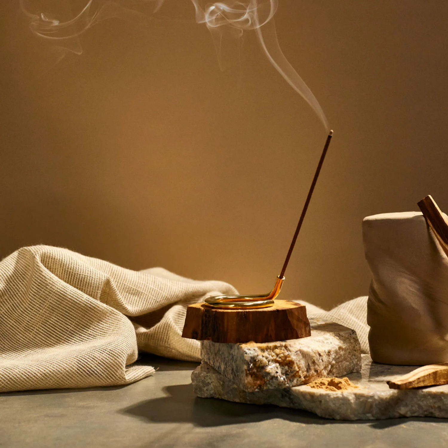 Palo Santi incense burned in a brass holder a top of some slabs of stone, wood and earthy linen fabric.