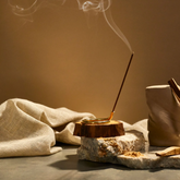 Palo Santi incense burned in a brass holder a top of some slabs of stone, wood and earthy linen fabric.