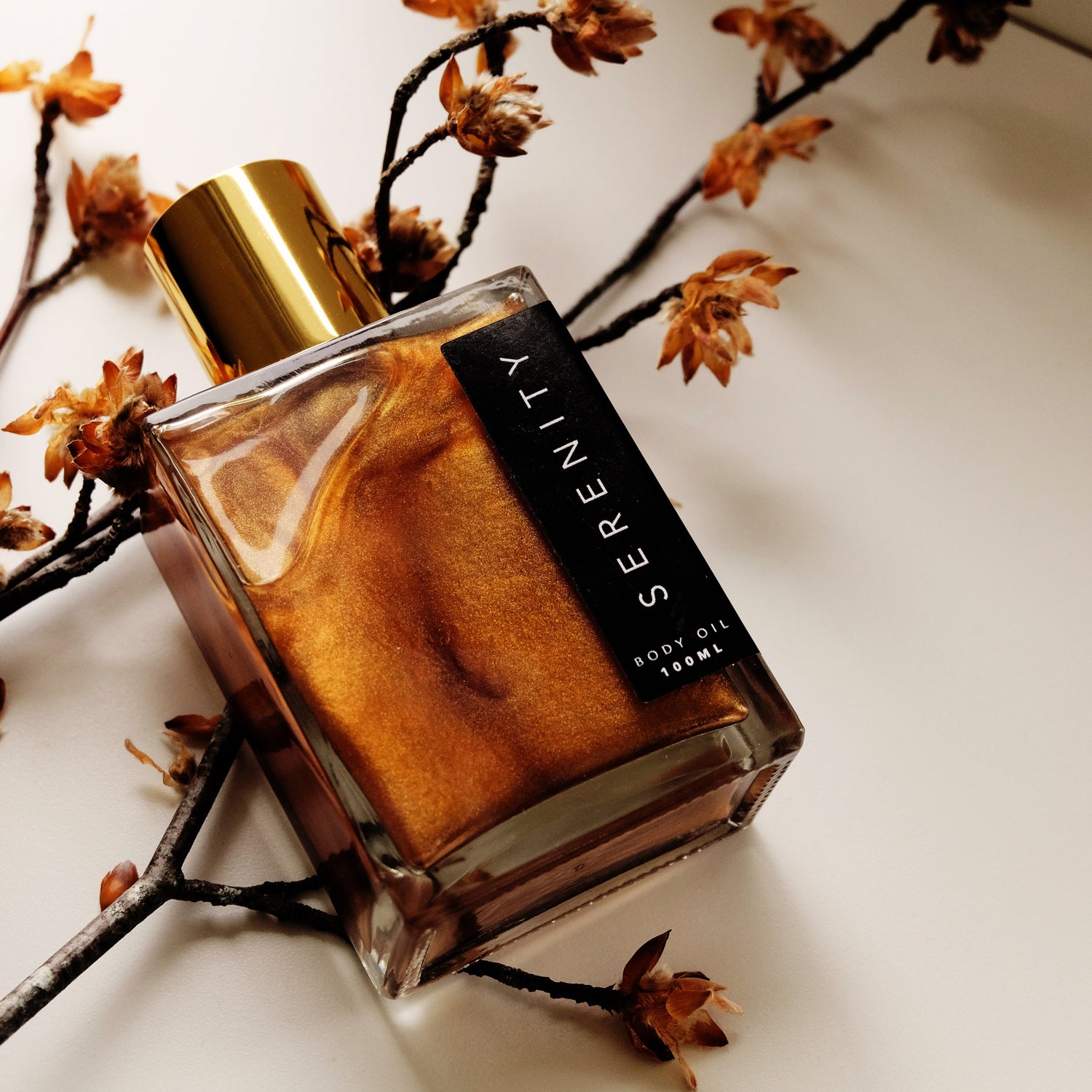 Square glass bottle containing shimmering golden body oil, lying on autumnal twigs.