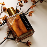Square glass bottle containing shimmering golden body oil, lying on autumnal twigs.