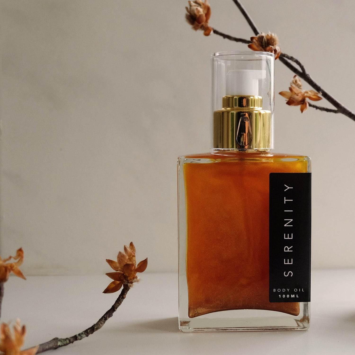 Square glass bottle containing golden body oil, soft white lighting with a couple of autumnal twigs.