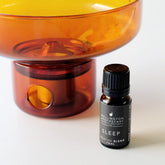 A black bottle of Sleep Sedative Oil Blend next to an amber glass oil burner.