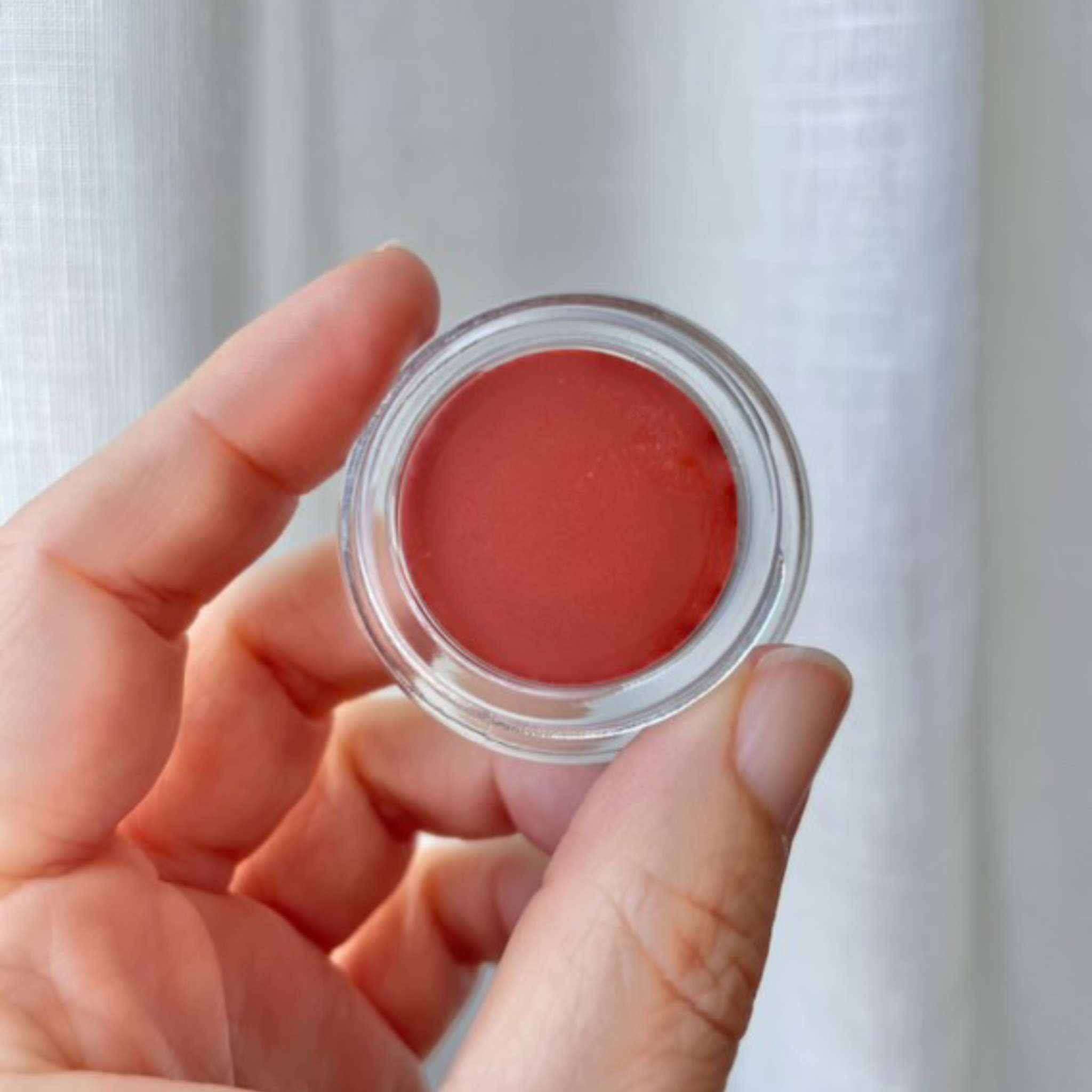 Hand holding a pottle of Terra lip tint in front of white linen curtain.
