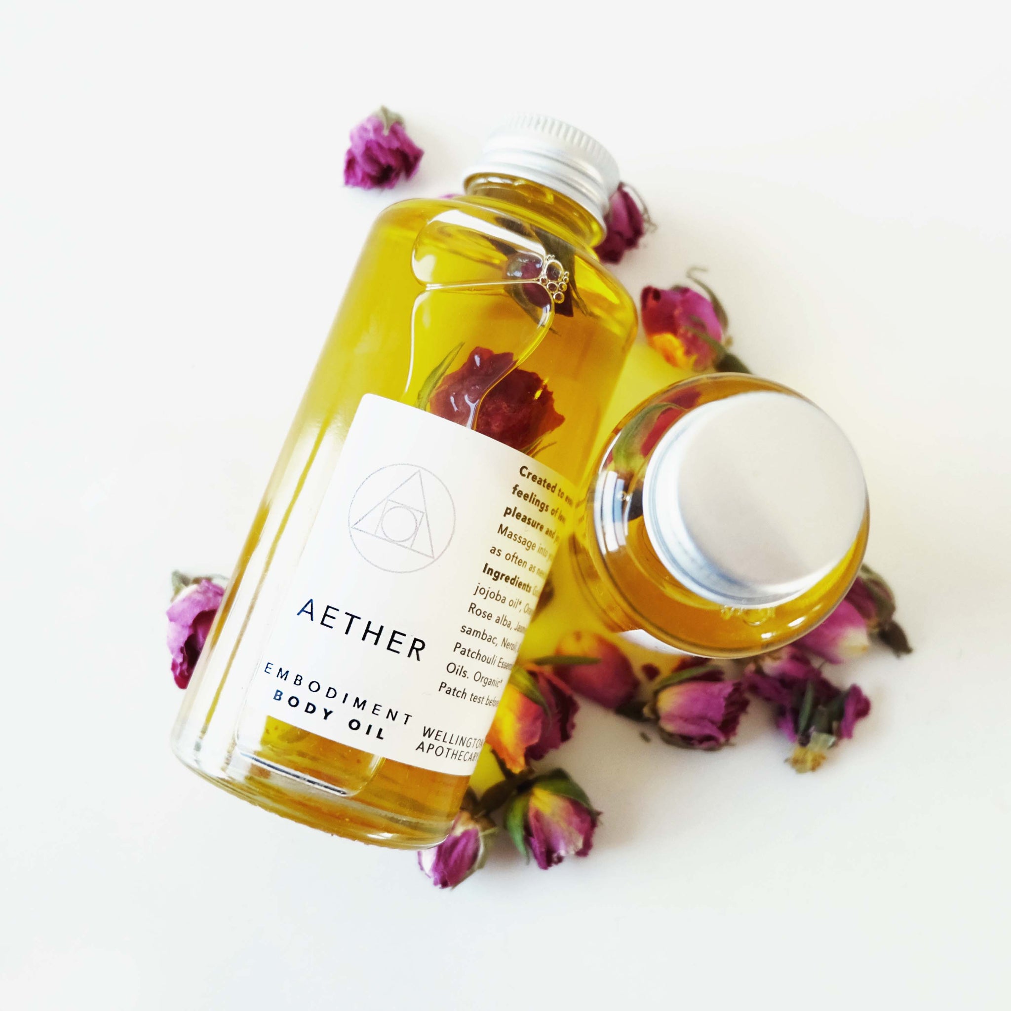 Clear glass bottles of golden Aether body oil infused with roses and surrounded by dried pink roses.