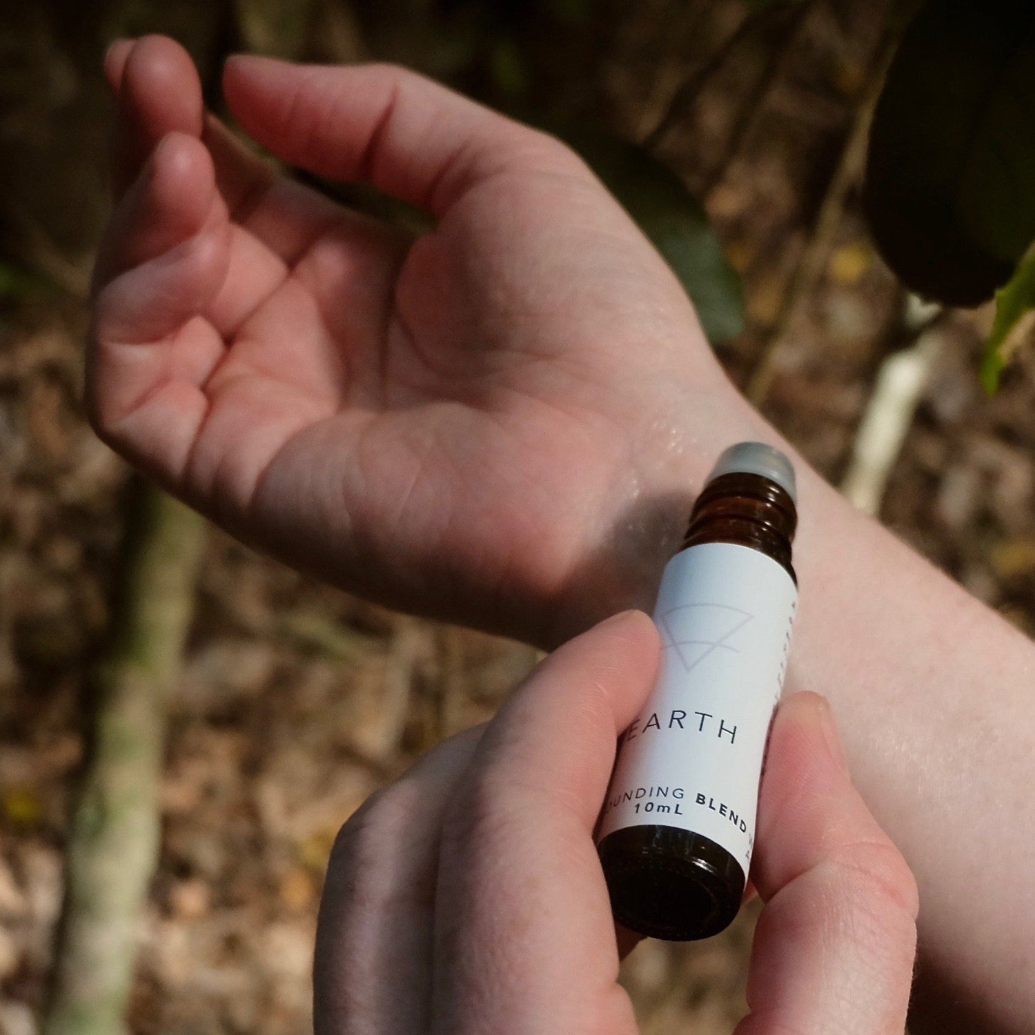 A small amber glass bottle of the Earth aromatherapy oil blend being rolled on a wrist in natural earthy surroundings.