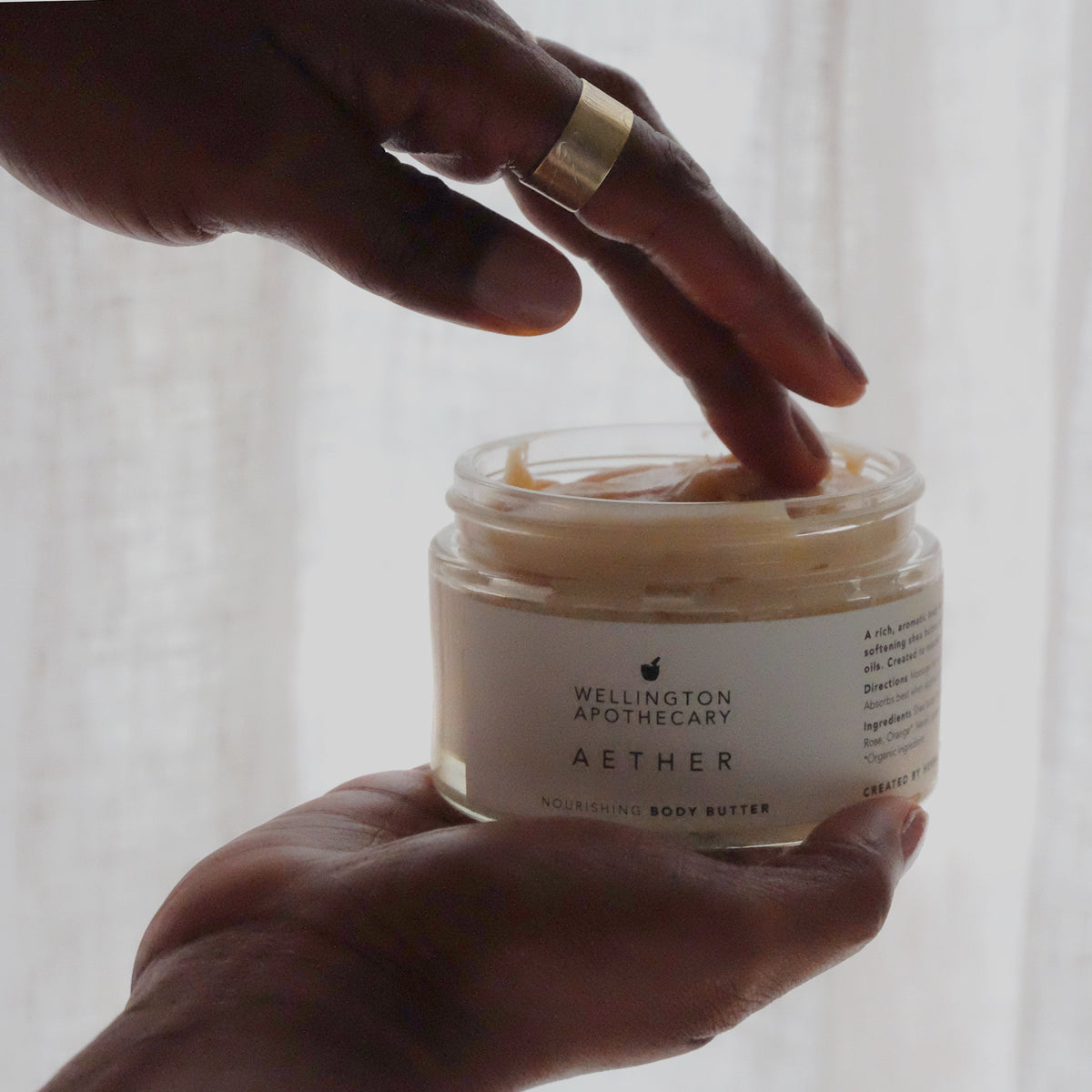 Woman's hands holding large glass jar of aether nourishing body butter, dipping a finger in, with soft light and sheer curtains as backdrop. 
