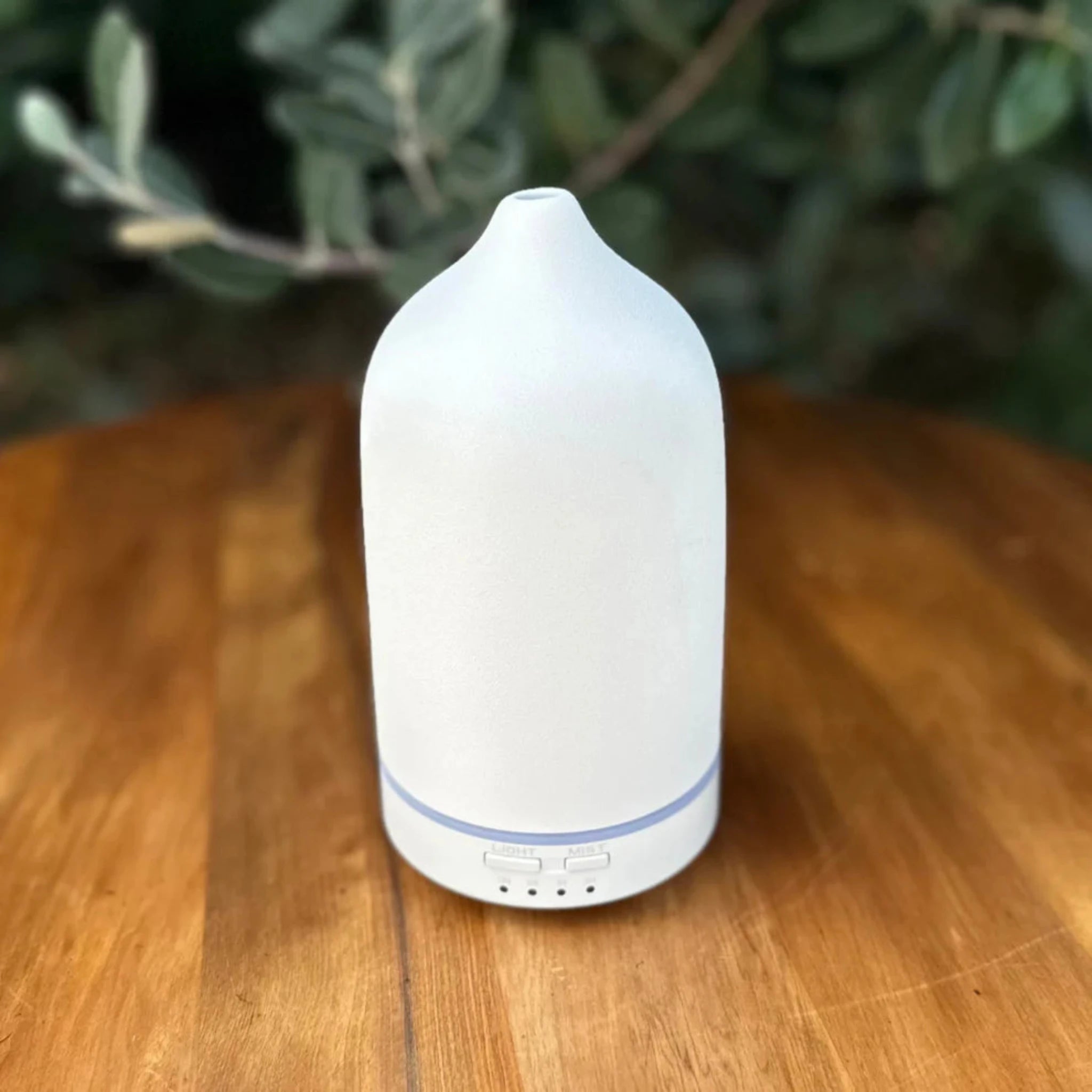 White essential oil diffuser on a wooden surface with greenery in the background