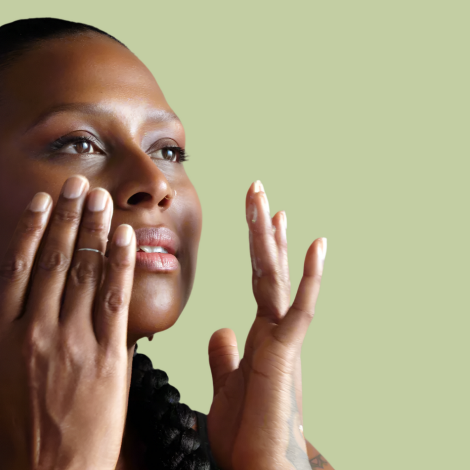Beautiful woman applying eye serum calmly while gazing into the distance with light green background.