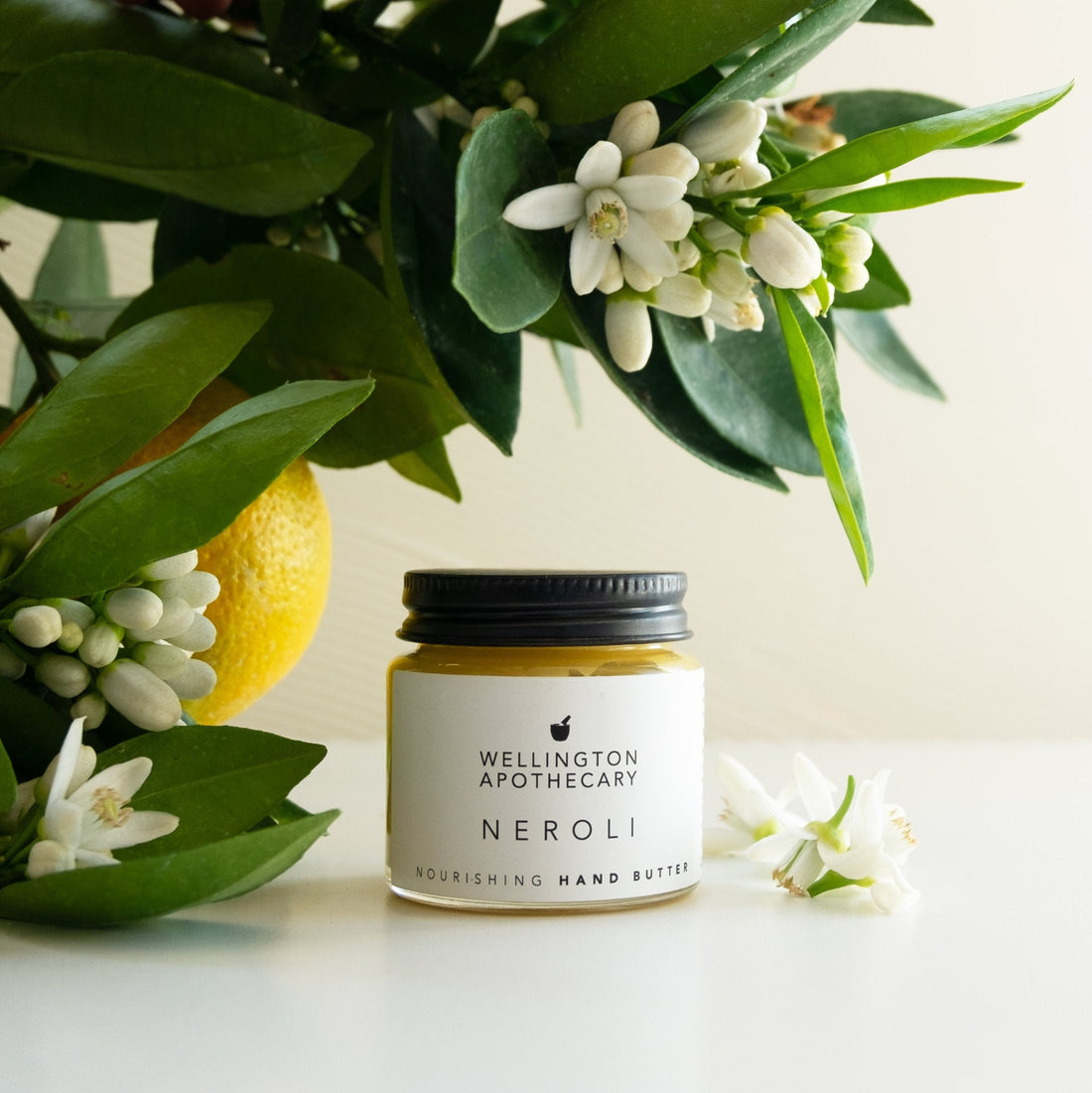 Glass jar of neroli hand butter on white bench, surrounded by orange blossoms, foliage and fruit. 