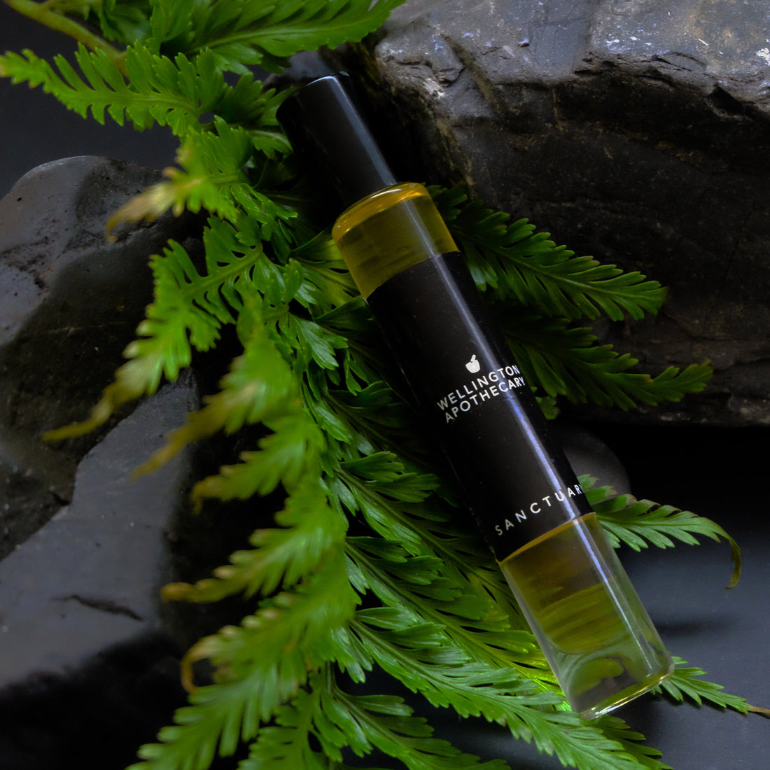 A clear glass roll on bottle containing Sanctuary perfume oil, resting on some rocks and a fern leaf. 