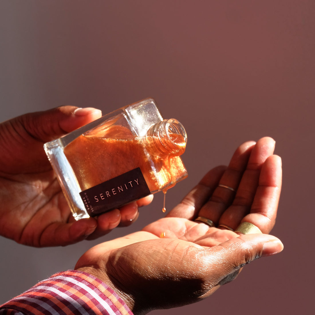Hand pouring shimmering body oil from a bottle labeled &