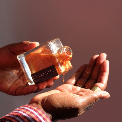 Hand pouring shimmering body oil from a bottle labeled &