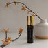 An amber glass vial of serenity perfume blend with gold lid, autumnal twigs and an earthy white ceramic vase surrounding. 