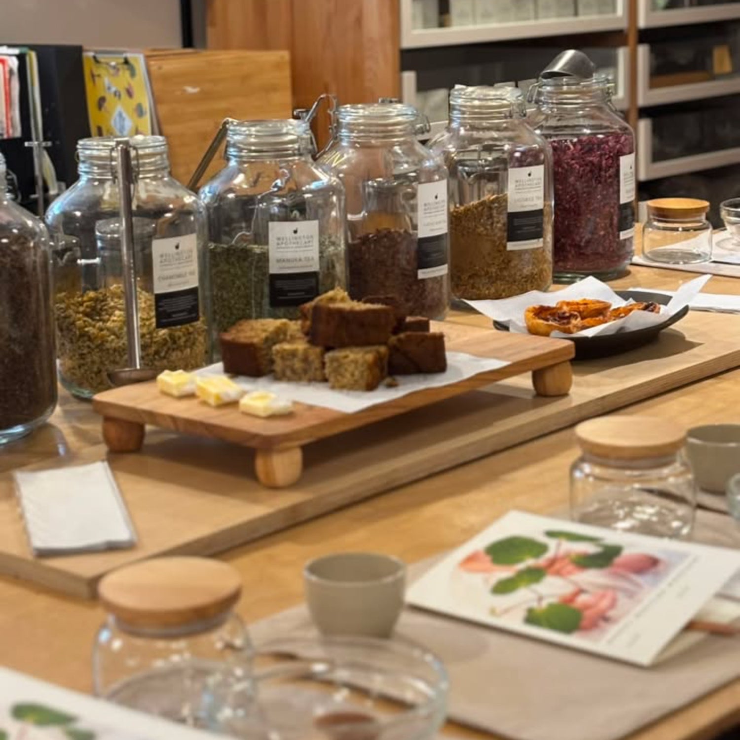 A styled tabletop display of glass jars filled with loose-leaf teas, herbs, and spices, alongside small cakes and butter on a wooden board in a kitchen or cafe setting.