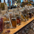 A wooden counter lined with clear glass jars containing loose-leaf teas, herbs, and spices, each with metal spoons resting inside. The background shows a softly blurred cafe interior with warm lighting and plants.