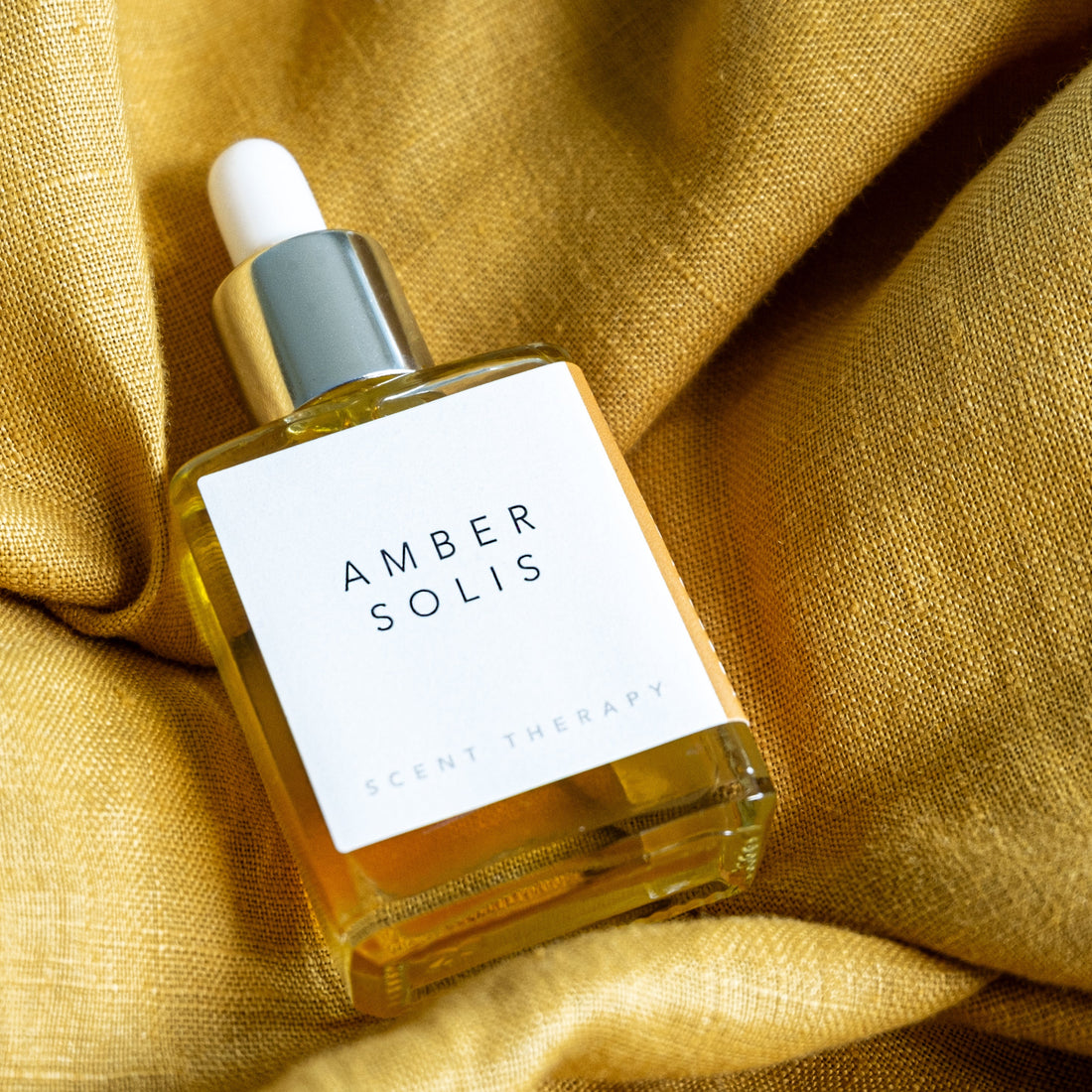 A square glass bottle of Amber Solis oil lying on ruffled golden chartreuse linen. 