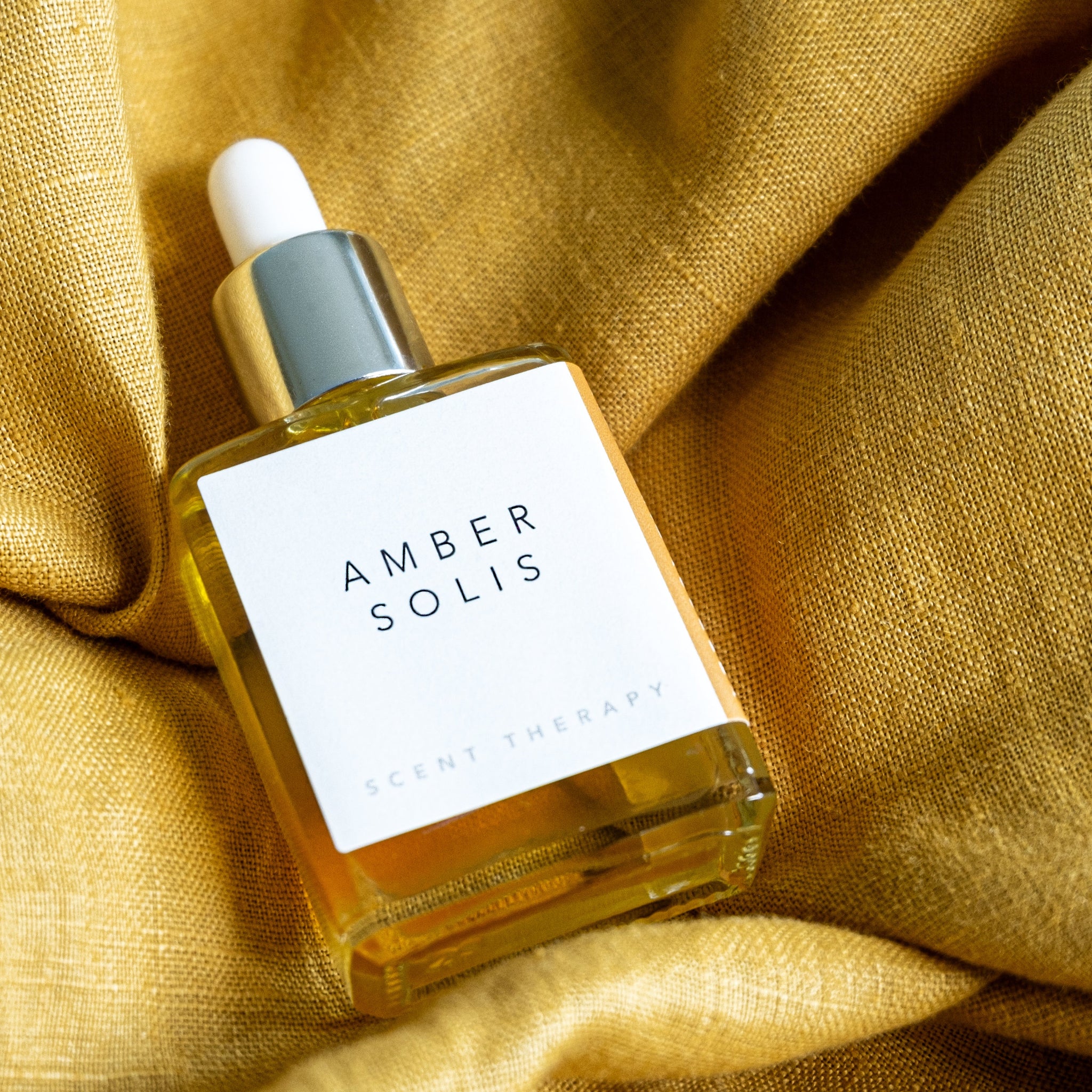 A square glass bottle of Amber Solis oil lying on ruffled golden chartreuse linen. 