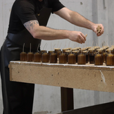 An industrial bench lined with amber glass candles being made by man in black apron.