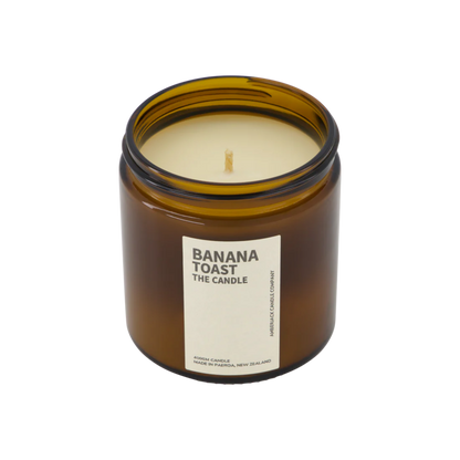 A large amber glass jar with brass lid containing banana toast candle on white background. 