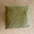 A square olive green linen throw pillow with a small white fabric tag on the side, resting on a light-colored wooden surface.
