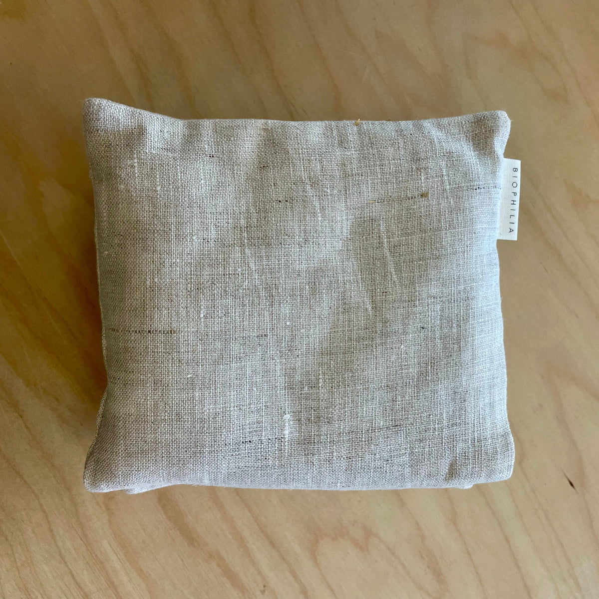 A square beige linen throw pillow with a small white fabric tag on the side, resting on a light wood grain surface.