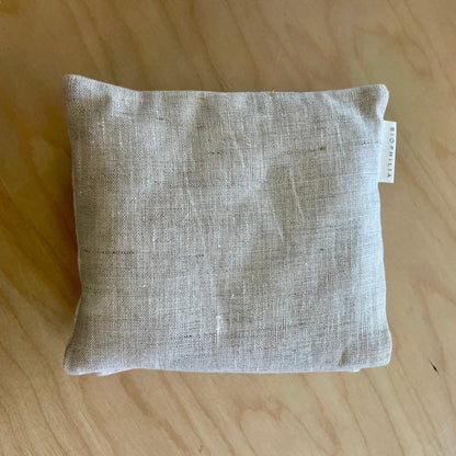 A square beige linen throw pillow with a small white fabric tag on the side, resting on a light wood grain surface.