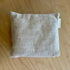 A square beige linen throw pillow with a small white fabric tag on the side, resting on a light wood grain surface.