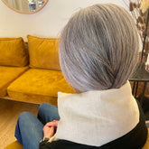 A person seated indoors, shown from behind, wearing a light beige linen neck pillow. The background features a mustard yellow velvet sofa and a neutral wall.