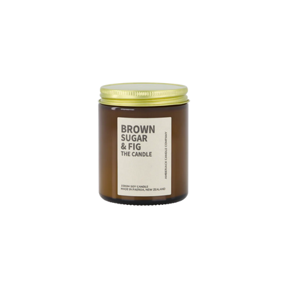 Amber glass jar with brass lid containing brown sugar and fig candle on white background. 