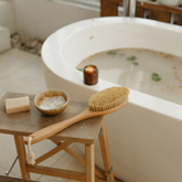 Bath setup with a wooden stool, brush, soap, and candle by a bathtub.