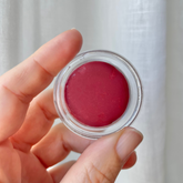 Hand holding an open pottle of Carmín lip tint in front of white linen curtain. 
