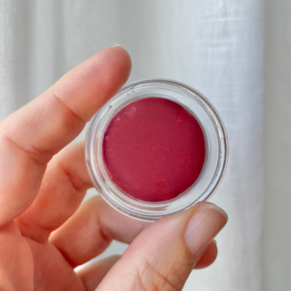 Hand holding an open pottle of Carmín lip tint in front of white linen curtain. 