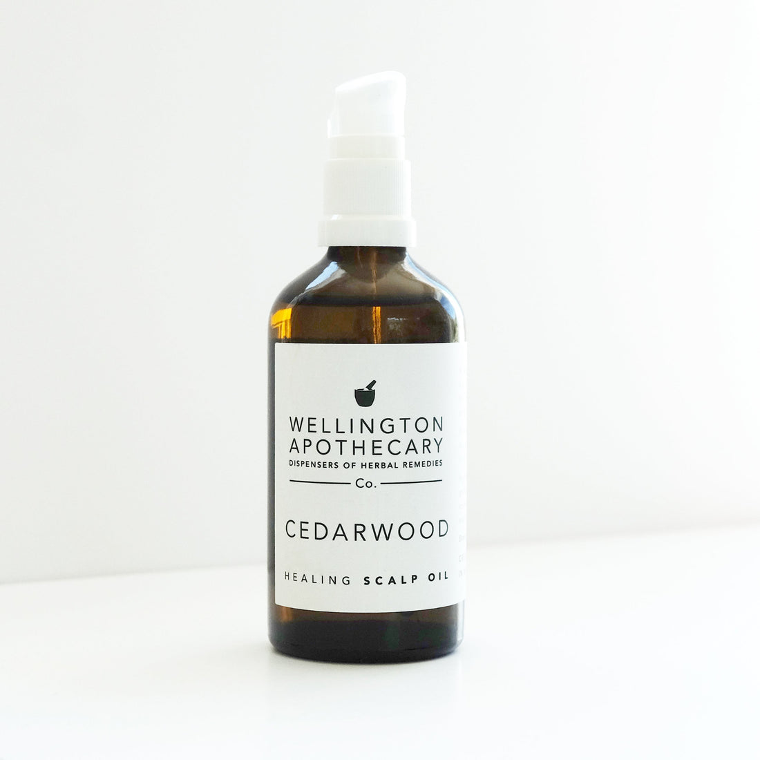 An amber glass bottle with a white pump top filled with Cedarwood Scalp Oil  on a white bench. 
