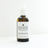 An amber glass bottle with a white pump top filled with Cedarwood Scalp Oil  on a white bench. 