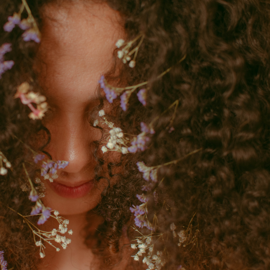Woman with curly brown hair decorated with white and purple dried flowers.